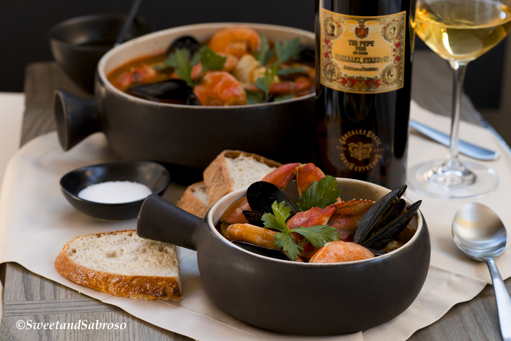 California-Coastal-Seafood-Stew- Served-In-Small-Grey-Bowl-With-Shrimp-Mussels-Dungeness-Crab-With-Sourdough-Bread-Bottle-of-Wine-and-Larger-Serving-Bowl-In-Background-With-Same-Ingredients
