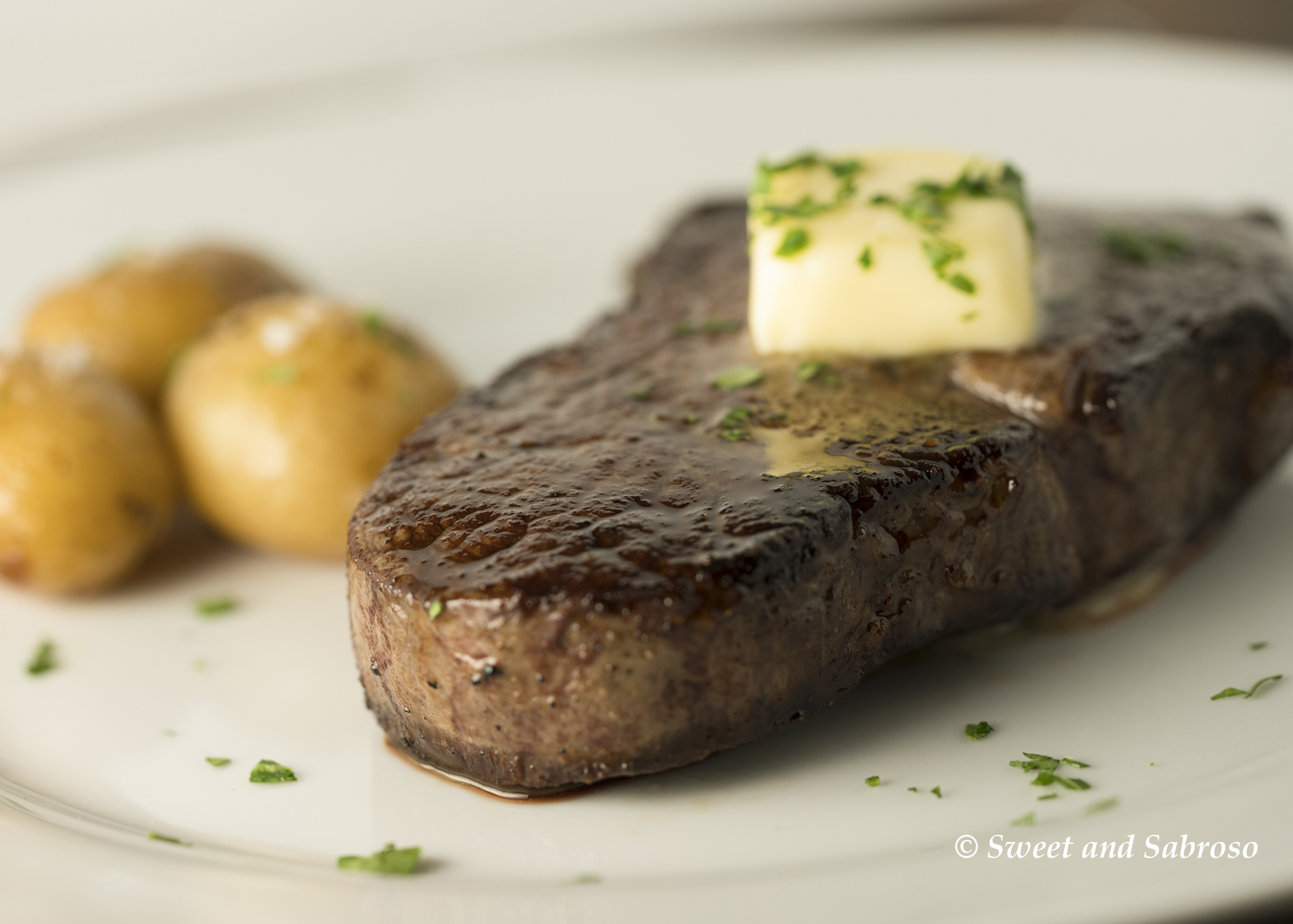 Perfect Pan Seared Restaurant Style Filet Mignon pictured with butter and parsley on top and two roasted potatoes in the background